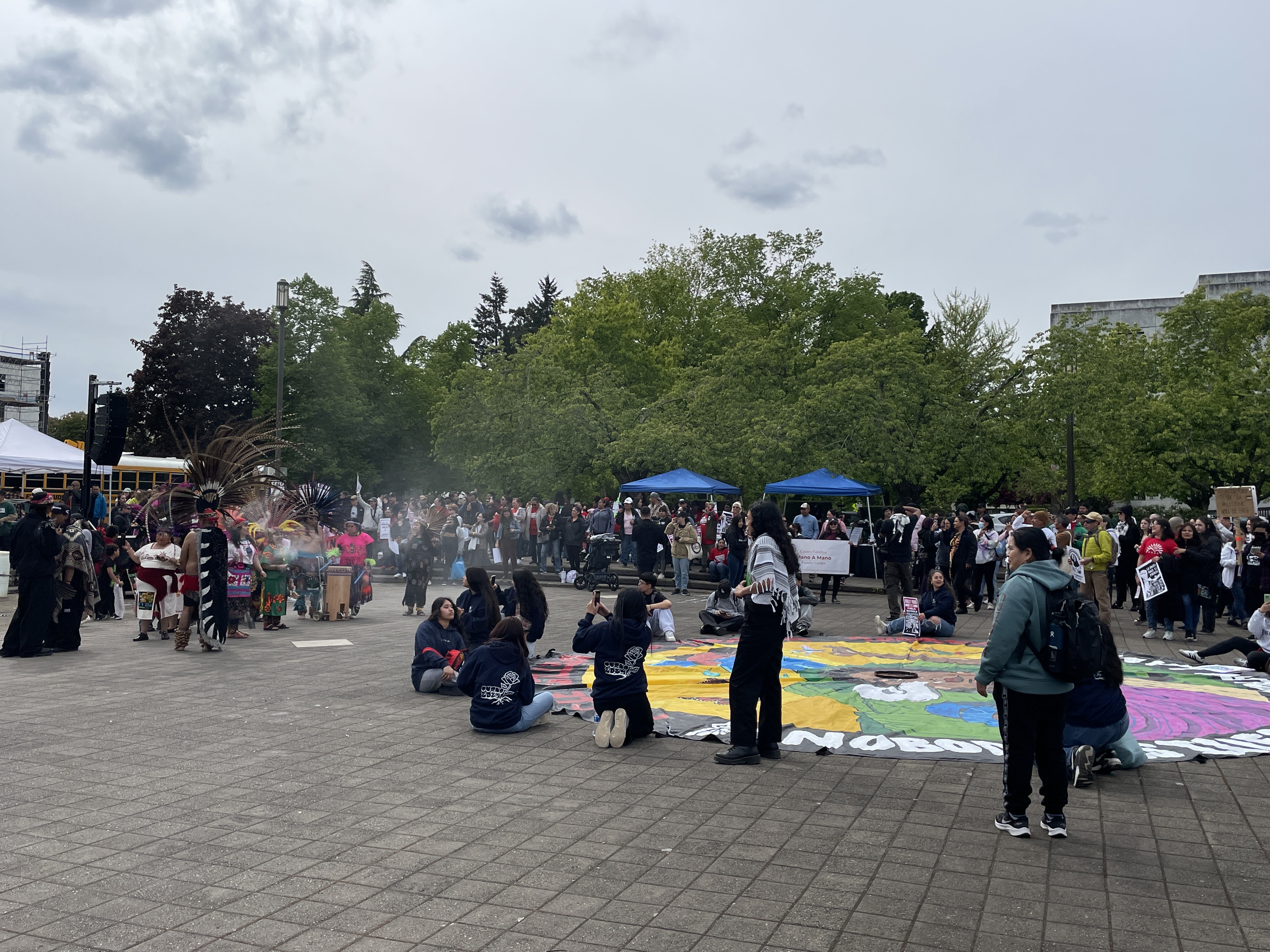 A large group of people gather outdoors on a paved area with trees in the background; some are seated or standing around a colorful circular artwork on the ground.
